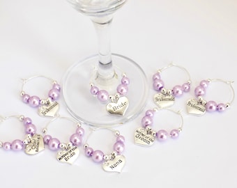 Könnte beinhalten: Satz mit sechs lila Glasanhängern mit silbernen Herzanhängern. Die Anhänger sind mit "Bride", "Chief Bridesmaid", "Mother of the Bride", "Mother of the Groom", "Bridesmaid" und "Nana" beschriftet.