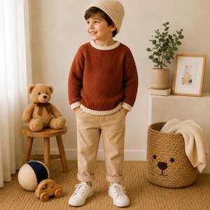 May include: A child wearing a textured rust-coloured long-sleeve jumper with cream-coloured ribbed trim at the neck, cuffs, and hem, paired with beige trousers and white trainers. The child also wears a cream-coloured knit beanie hat.