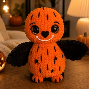 May include: A plush toy shaped like a round orange pumpkin creature with large black glittery eyes, a small black heart-shaped nose, and a white stitched smile. It features black wings, black star-shaped accents on its body, and small orange feet.