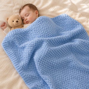 Può includere: Una coperta per neonati in maglia azzurra con un motivo a nido d'ape testurizzato e un bordo a coste. La coperta copre un neonato addormentato che tiene in mano un piccolo orsacchiotto di peluche marrone chiaro con occhi scuri e naso ricamato.