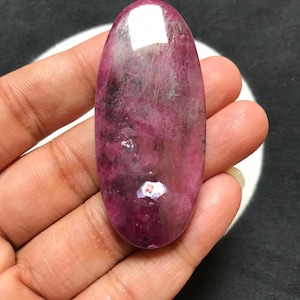 Könnte beinhalten: Ein oval geschliffener Edelstein in tiefen Magenta- und Rosatönen mit natürlichen Einschlüssen, dunklen Mineralflecken und einer glatten, reflektierenden Oberfläche, die das Licht über die längliche, abgerundete Form einfängt.