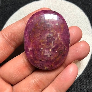 May include: An oval-shaped polished gemstone with a deep magenta and purple base colour, featuring mottled inclusions of white, yellow, and black mineral flecks throughout the smooth, glossy surface of the stone.