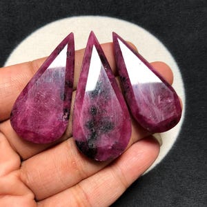 以下が含まれることがあります： 深いマゼンタとピンクのカラーパレットに、暗い黒のインクルージョンと磨かれた反射面を持つ、3つの涙型のカボション。各石は滑らかでファセット加工された上端と、自然な繊維状の内部テクスチャを示しています。