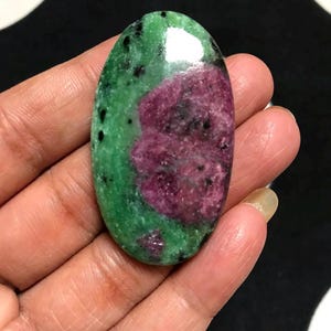 Puede incluir: Una piedra preciosa pulida de forma ovalada con un color base verde vibrante, inclusiones prominentes de color magenta rosado y motas negras dispersas por toda la superficie. La piedra tiene un acabado liso y brillante que refleja la luz.