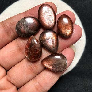 Puede incluir: Una colección de seis cabujones de piedra de sol pulidos en varias formas, incluyendo óvalos y gotas. Estas piedras exhiben una aventurescencia metálica brillante con tonos rojo-marrones profundos, cobre y gris oscuro en sus superficies.