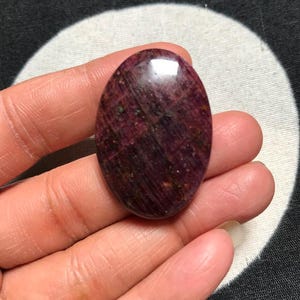 Peut inclure: Une pierre précieuse cabochon de forme ovale avec une palette de couleurs bordeaux profond et rouge foncé. La pierre présente une surface lisse et polie avec des inclusions naturelles visibles et un éclat subtil qui reflète la lumière.