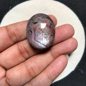 Puede incluir: Una piedra preciosa pulida de forma ovalada que presenta una mezcla de tonos morados profundos, marrón rojizo y gris con inclusiones negras visibles y una superficie lisa y reflectante que capta la luz, mostrando patrones minerales naturales.
