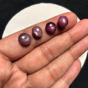 Peut inclure: Quatre petites pierres précieuses rondes taillées en cabochon avec une teinte rouge-violet profond. Chaque pierre présente une surface lisse et polie avec un léger reflet lumineux en forme d'étoile, appelé astérisme, sur le dôme.