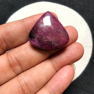 May include: A polished, triangular-shaped gemstone with a smooth, domed surface. The stone features a deep magenta and dark purple colour palette with visible natural inclusions and a reflective sheen across its top portion.