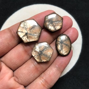 May include: Four polished gemstones with a light brown, translucent appearance and dark, intersecting linear inclusions. Two stones are hexagonal, one is smaller and hexagonal, and one is oval, all displaying a distinct star-like pattern.