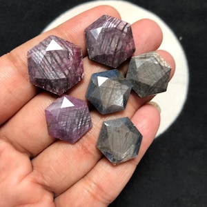May include: Five loose, hexagonal-cut gemstones with a rose-cut top facet. Three stones display deep purple and magenta hues with visible internal striations, while two stones exhibit muted grey and blue tones with similar linear inclusions.