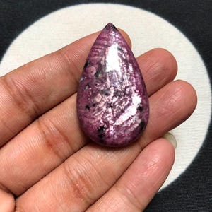 Könnte beinhalten: Ein tropfenförmiger Cabochon-Edelstein mit einer tief magentafarbenen und violetten Grundfarbe, markanten schwarzen Mineraleinschlüssen und einer polierten, reflektierenden Oberfläche, die das Licht auf der kristallinen Struktur einfängt.