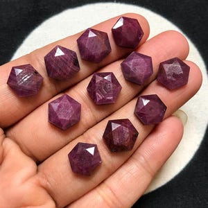 Pode incluir: Uma coleção de dez rubis soltos com corte hexagonal em uma cor vermelho-magenta profunda e saturada. Cada pedra preciosa apresenta uma superfície facetada que reflete a luz, exibindo uma forma geométrica consistente e um aspecto polido.