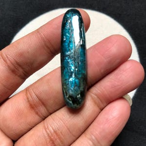 Puede incluir: Una piedra preciosa pulida de forma ovalada alargada con un color azul verdoso vibrante y profundo y una textura superficial cristalina y reflectante. La piedra presenta inclusiones internas y variaciones naturales en su composición.