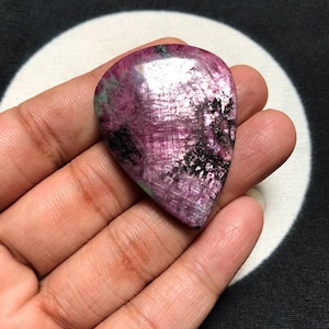 Pode incluir: Uma pedra preciosa polida em forma de lágrima com uma cor base vibrante em magenta e rosa profundo, apresentando inclusões minerais pretas proeminentes por toda a superfície. A pedra tem um acabamento liso e reflexivo com textura natural.
