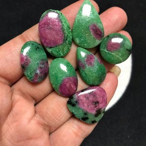 Puede incluir: Una colección de siete piedras preciosas pulidas de rubí en zoisita con bases minerales de color verde vibrante e inclusiones de rubí magenta-rosa. Las piedras tienen formas variadas de óvalos lisos, gotas y formas redondeadas.