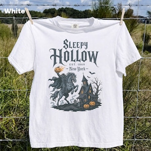 May include: A white t-shirt featuring a graphic print with the text Sleepy Hollow Est. 1869 New York. The illustration depicts a headless horseman riding a horse while holding a flaming pumpkin, a dark house, bare trees, and two orange pumpkins.