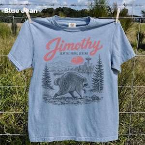 May include: A light blue t-shirt featuring a graphic print of a raccoon in the foreground with a Seattle cityscape, a red sun, and evergreen trees in the background. The text reads Jimothy Seattle Feral Legend in a red script font.
