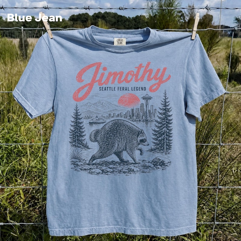May include: A light blue t-shirt featuring a graphic print of a raccoon in the foreground with a Seattle cityscape, a red sun, and evergreen trees in the background. The text reads Jimothy Seattle Feral Legend in a red script font.