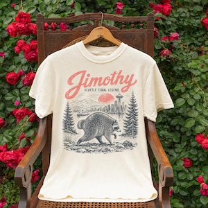 May include: A cream-colored t-shirt featuring a graphic print of a raccoon walking in a forest with a city skyline and the Space Needle in the background. The text reads "Jimothy" in red script and "SEATTLE FERAL LEGEND" in black block letters.