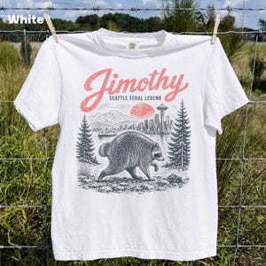 May include: A white short-sleeve t-shirt featuring a graphic print of a raccoon walking in a forest. The text reads Jimothy Seattle Feral Legend in pink and black ink. The design includes a mountain range, a city skyline, and a red sun.