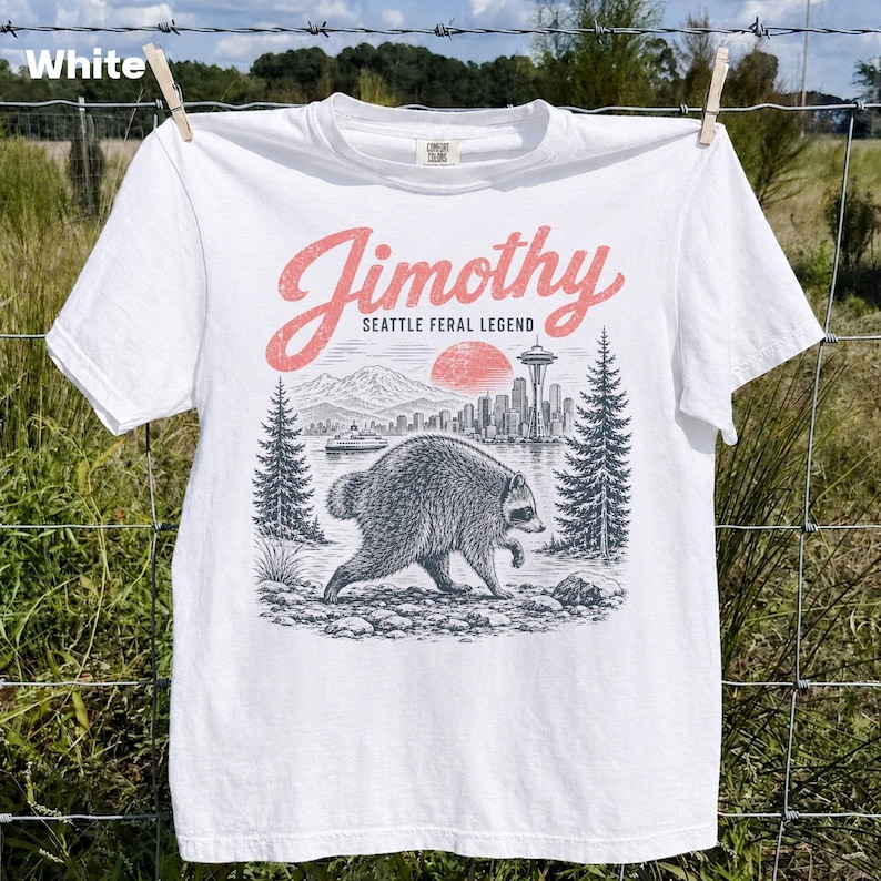 May include: A white short-sleeve t-shirt featuring a graphic print of a raccoon walking in a forest. The text reads Jimothy Seattle Feral Legend in pink and black ink. The design includes a mountain range, a city skyline, and a red sun.