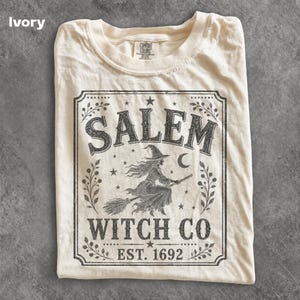May include: An ivory t-shirt featuring a dark gray graphic print of a witch flying on a broomstick. The design is enclosed in a rectangular border with the text SALEM WITCH CO EST. 1692, surrounded by stars, crescent moon, and leaf motifs.