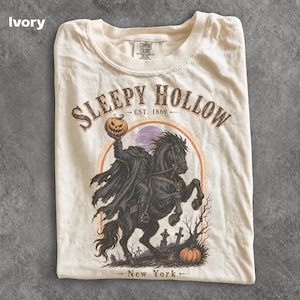 May include: An ivory t-shirt featuring a graphic of the Headless Horseman riding a black horse, holding a pumpkin. The text reads SLEEPY HOLLOW EST. 1869 NEW YORK. The design includes a purple moon, bare trees, and tombstones in a circular frame.
