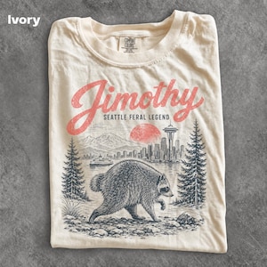 May include: An ivory t-shirt featuring a graphic print of a raccoon in the foreground with a Seattle cityscape, the Space Needle, a ferry, and mountains in the background. The text reads Jimothy SEATTLE FERAL LEGEND in red and black ink.