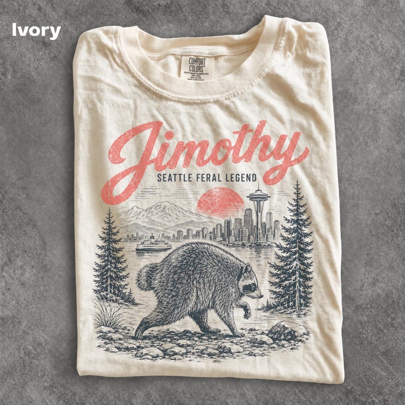 May include: An ivory t-shirt featuring a graphic print of a raccoon in the foreground with a Seattle cityscape, the Space Needle, a ferry, and mountains in the background. The text reads Jimothy SEATTLE FERAL LEGEND in red and black ink.