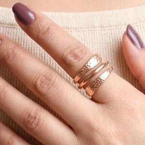 Könnte beinhalten: Ein Set aus drei Ringen in Roségold, die an einem Finger getragen werden. Das Set besteht aus einem Ring mit gehämmerter Textur, einem glatten, dünnen Band und einem Ring mit texturierter, genoppter Oberfläche. Die Ringe sind gestapelt.