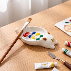 May include: A white ceramic cat-shaped paint palette with small dabs of red, blue, yellow, and green oil paint. A wooden-handled paintbrush rests across the palette, accompanied by a metal palette knife and tubes of paint nearby.