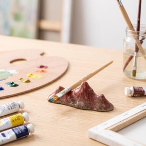 May include: A ceramic brush rest shaped like a mountain with a mottled red and green glaze. The brush rest holds a single wooden paintbrush with a light-coloured handle and a metal ferrule. The object is positioned on a flat surface.