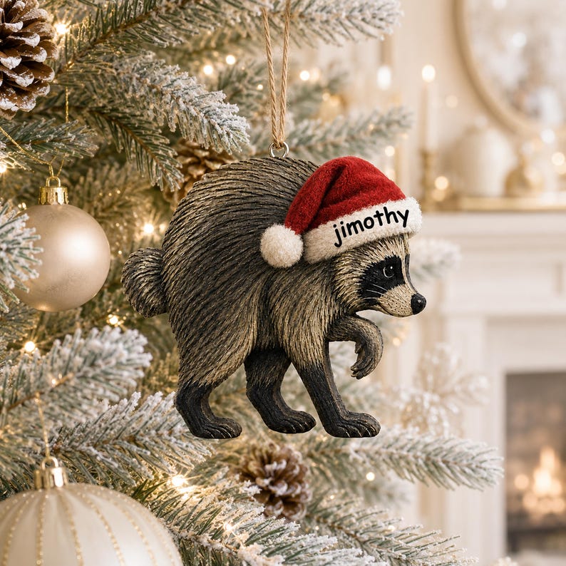 May include: A raccoon Christmas tree ornament with textured brown and black fur, wearing a red and white Santa hat with the name Jimothy printed in black on the white brim. The ornament hangs from a thin brown twine loop against a pine tree.