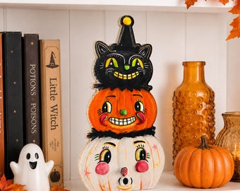 May include: A vertical wooden cutout decoration featuring a stack of three characters: a black cat in a pointed hat on top, an orange jack-o'-lantern in the middle, and a white pumpkin face at the base, all with painted vintage-style facial features.