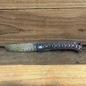 Peut inclure: Un couteau de poche pliant doté d'une lame en acier damas au motif tourbillonnant et d'un manche en matériau composite multicouche. Le point de pivot est fixé par une vis métallique bleu vif et une goupille de couleur argentée.