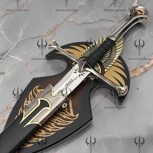 Puede incluir: Una espada de metal plateado con empuñadura de cuero negro y una guarda con detalles dorados. La hoja está grabada con caracteres rúnicos. Está montada sobre una placa de madera negra decorada con motivos de alas doradas.