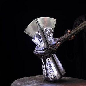 May include: A large silver hammer with a metallic head and a dark, textured, branch-like handle. The head features glowing white lightning patterns that illuminate the surface. The design includes a wide, flat top and a rectangular base.