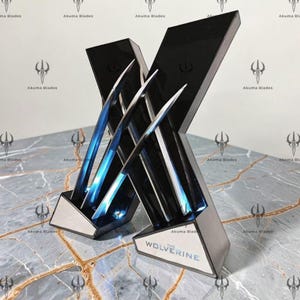 May include: A black display stand shaped like an X, holding three metallic claw blades with blue LED lighting. The base features the text THE WOLVERINE in black lettering. The structure is angled, showcasing sharp, pointed edges and a sleek finish.