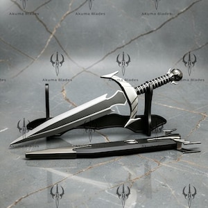 May include: A silver and matte black dagger with a pointed blade, a curved crossguard, and a ribbed handle with a spherical pommel, resting on a black stand. A matching black and silver scabbard lies in front, featuring the text Akuma Blades.