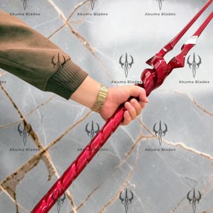 May include: A long, metallic red spear featuring a spiraled shaft and a multi-pronged, pointed head. The weapon has a glossy finish and is held by a hand wearing a gold-toned metal link watch, with the text Akuma Blades visible in the background.
