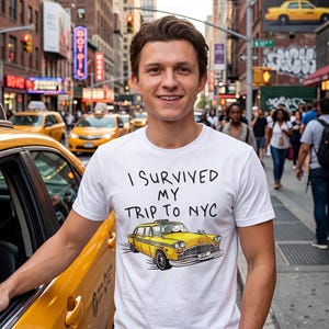 May include: A white short-sleeved t-shirt featuring the black text I SURVIVED MY TRIP TO NYC above a graphic illustration of a yellow taxi cab with a cartoon character in the driver seat, set against a busy New York City street background.