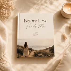 May include: A hardback book with a cream-coloured cover featuring the title Before Love Finds Me in dark brown text, a small line drawing of a sprig, and a photograph of a person in a white dress walking on a path towards the ocean.