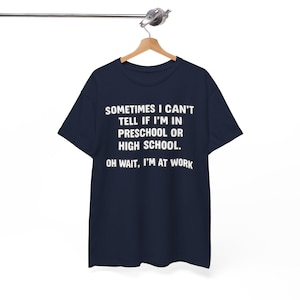 May include: A dark navy blue short-sleeved t-shirt featuring white text that reads: SOMETIMES I CAN'T TELL IF I'M IN PRESCHOOL OR HIGH SCHOOL. OH WAIT, I'M AT WORK. The shirt is displayed on a wooden hanger suspended from a metal rod.