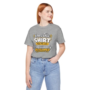 May include: A heather grey short-sleeved t-shirt featuring the text Does This SHIRT Make Me Look Professionally DEVELOPED in a mix of white and yellow stylised lettering. The shirt is paired with light blue denim jeans with a button fly closure.