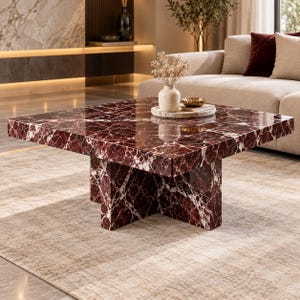 Puede incluir: Una mesa de centro cuadrada fabricada en mármol rojo intenso con vetas blancas prominentes. La mesa cuenta con una superficie gruesa y pulida, apoyada sobre una base robusta en forma de cruz hecha del mismo material de mármol rojo.