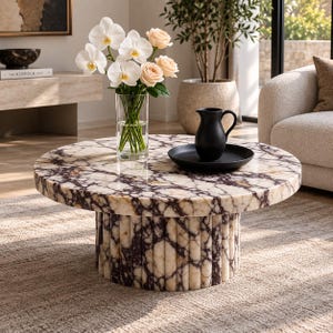 Op de afbeelding: Een ronde salontafel met een wit en donkerpaars marmeren blad en een bijpassende gecanneleerde cilindrische voet. Op het tafelblad staat een doorzichtige glazen vaas gevuld met witte orchideeën en lichtroze rozen, naast een zwart dienblad met een kan.