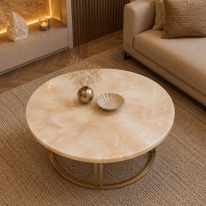 May include: A round coffee table featuring a smooth, light beige marble-patterned top with subtle white veining. The table is supported by a circular, thin gold-toned metal frame base, providing a stable and open structure for the tabletop surface.
