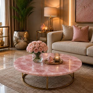 May include: A round coffee table featuring a polished pink rose quartz stone top supported by a circular gold metal frame. A clear glass vase filled with light pink roses and a small gold tray holding a candle and a crystal sit on the table surface.