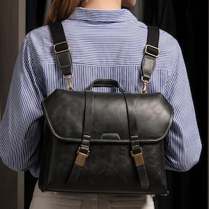 May include: A black leather-style backpack with a fold-over flap, two vertical straps with metallic buckles, and adjustable shoulder straps. The bag features a top handle and is worn over a light blue and white striped button-down shirt and white pants.